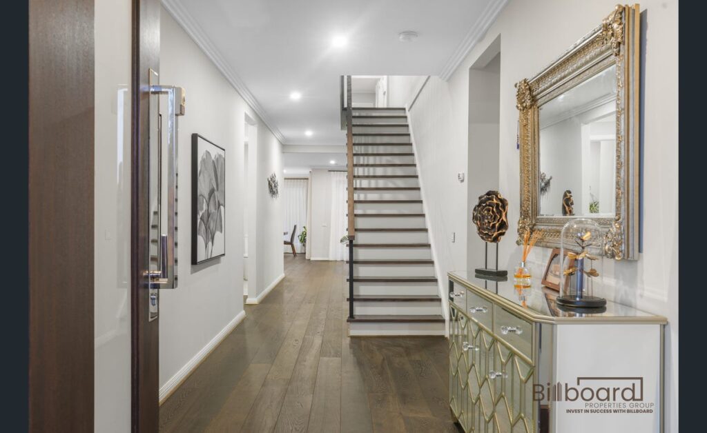 Elegant entrance hallway with dark timber flooring, decorative mirrored console table and gold framed mirror, modern staircase leading to upper level, framed wall art and bright recessed lighting enhancing the luxury interior feel.
