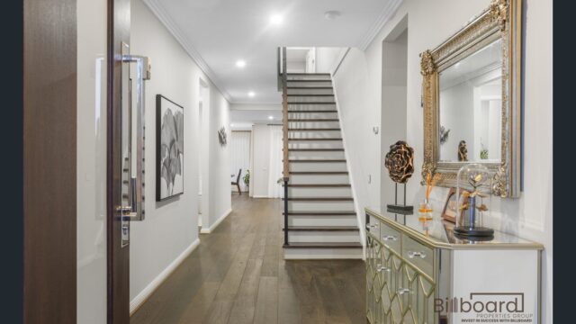 Elegant entrance hallway with dark timber flooring, decorative mirrored console table and gold framed mirror, modern staircase leading to upper level, framed wall art and bright recessed lighting enhancing the luxury interior feel.