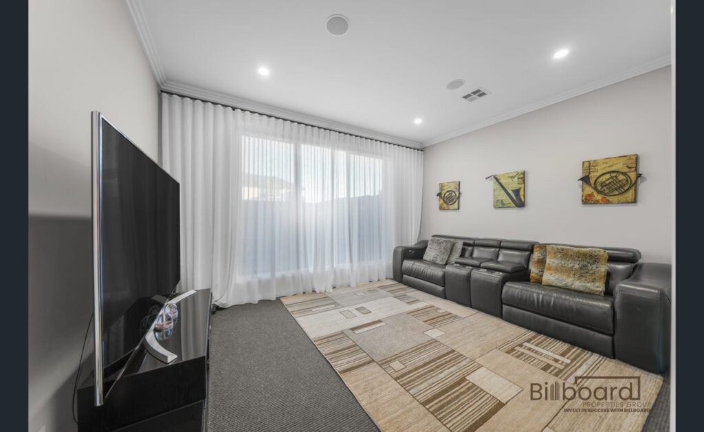 Cozy media room with a large flat-screen TV, black leather recliner sofa, patterned area rug, soft sheer curtains over wide windows, and decorative wall art creating a comfortable entertainment space.