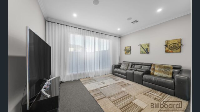 Cozy media room with a large flat-screen TV, black leather recliner sofa, patterned area rug, soft sheer curtains over wide windows, and decorative wall art creating a comfortable entertainment space.