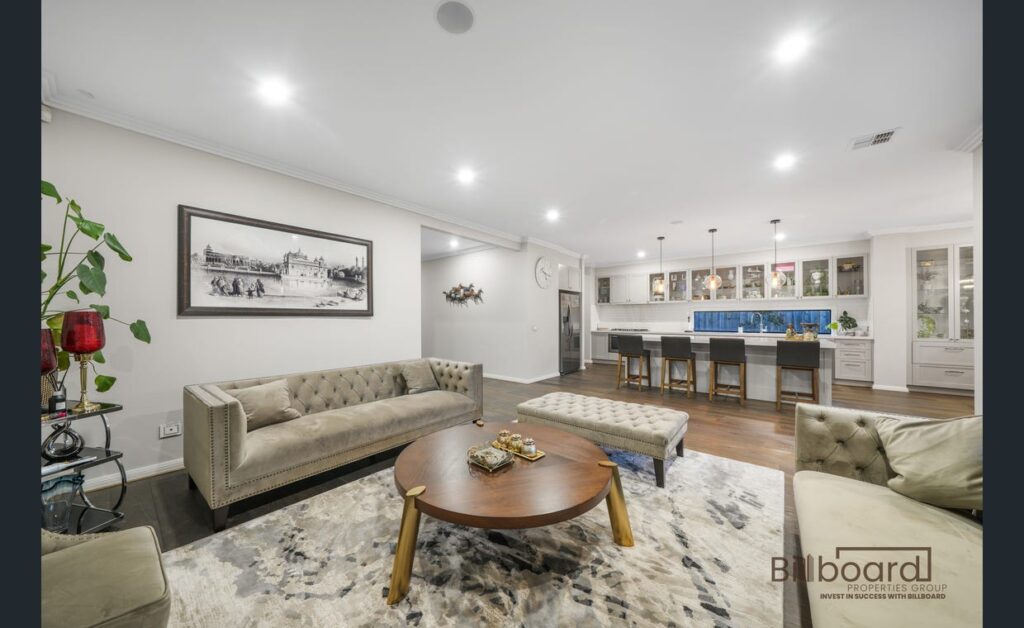 Spacious open-plan living area featuring tufted sofas, round wooden coffee table, decorative rug, indoor plants and wall art, with the kitchen visible in the background including bar stools, pendant lighting and ample cabinetry, creating a warm and luxurious family space.