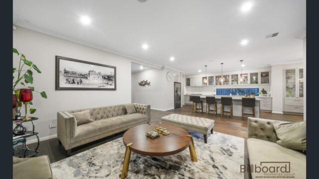 Spacious open-plan living area featuring tufted sofas, round wooden coffee table, decorative rug, indoor plants and wall art, with the kitchen visible in the background including bar stools, pendant lighting and ample cabinetry, creating a warm and luxurious family space.