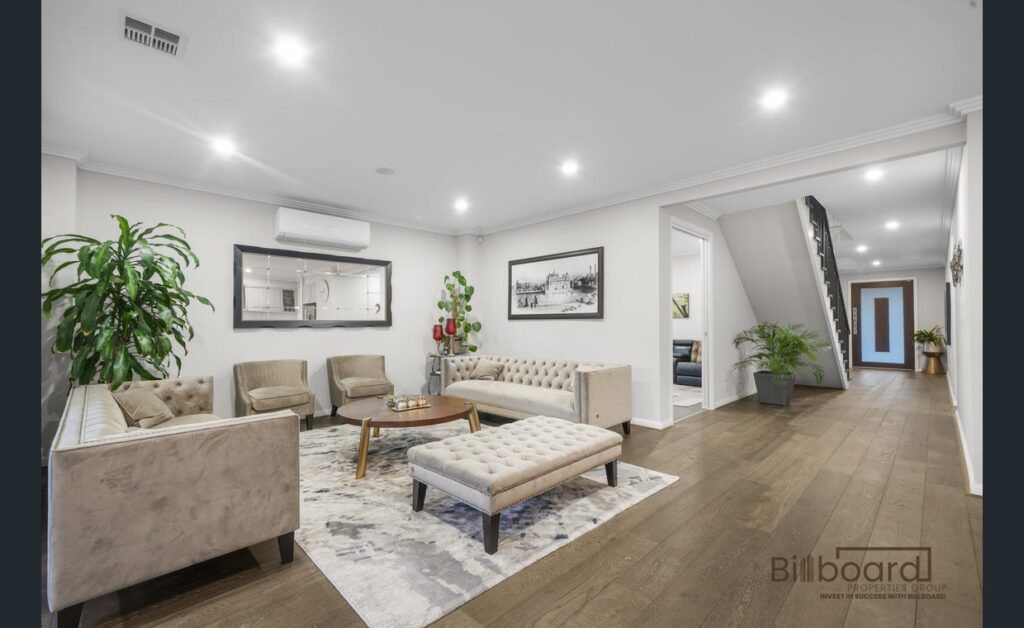 Elegant living room with tufted sofas and armchairs, ottoman-style coffee table, indoor plants, framed artwork on the wall, wooden flooring, and recessed ceiling lights, with a hallway and staircase visible leading to the rest of the home.