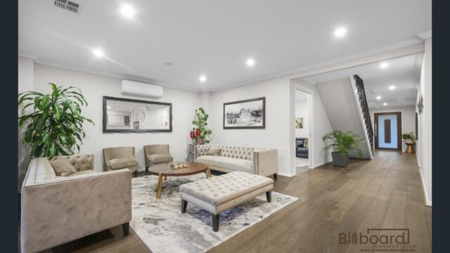 Elegant living room with tufted sofas and armchairs, ottoman-style coffee table, indoor plants, framed artwork on the wall, wooden flooring, and recessed ceiling lights, with a hallway and staircase visible leading to the rest of the home.