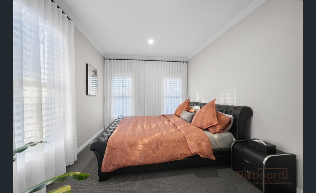 Bedroom with a black upholstered bed frame, peach-colored bedding, bedside table with decor, grey carpet flooring, and sheer white curtains covering windows along the walls, creating a soft and cozy atmosphere.