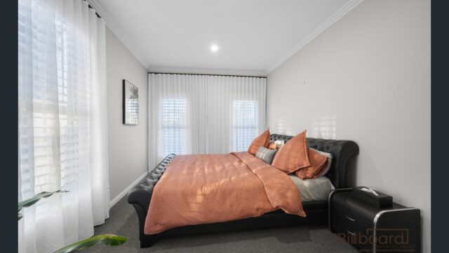 Bedroom with a black upholstered bed frame, peach-colored bedding, bedside table with decor, grey carpet flooring, and sheer white curtains covering windows along the walls, creating a soft and cozy atmosphere.