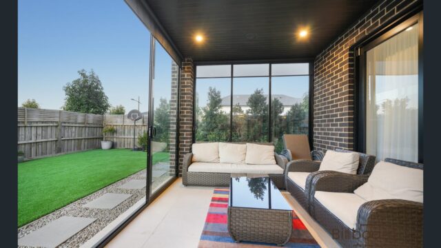Enclosed outdoor sitting area with wicker-style lounge furniture, glass sliding doors opening to a backyard with artificial grass, and cozy indoor-outdoor ambience under a dark ceiling with recessed lighting.