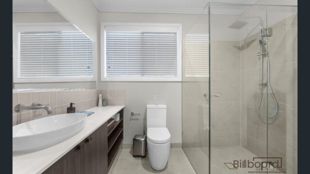 Modern bathroom with a glass walk-in shower, white toilet, floating vanity with vessel sink, large mirror, tiled walls and flooring, and two windows with blinds allowing natural light in.