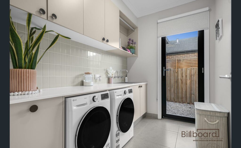 Modern laundry room with front-loading washing machine and dryer, long benchtop, overhead cabinetry, indoor plant decor, tiled flooring, and a door leading to the backyard area.