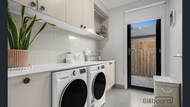 Modern laundry room with front-loading washing machine and dryer, long benchtop, overhead cabinetry, indoor plant decor, tiled flooring, and a door leading to the backyard area.