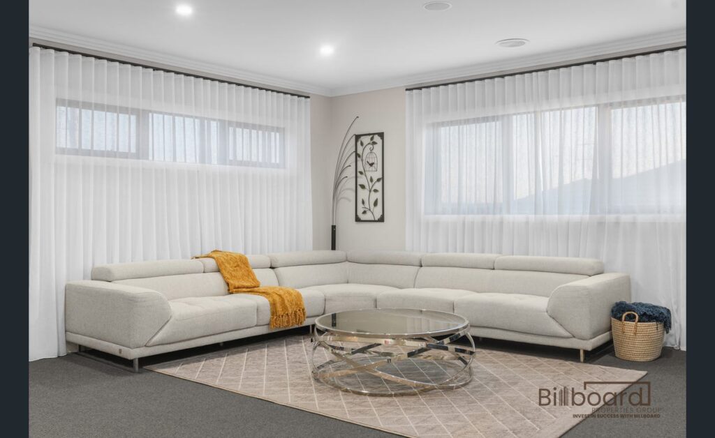 Stylish lounge area with a large cream L-shaped sofa, golden throw blanket, modern glass round coffee table, neutral decor accents, and sheer white curtains covering wide windows for a bright and contemporary look.
