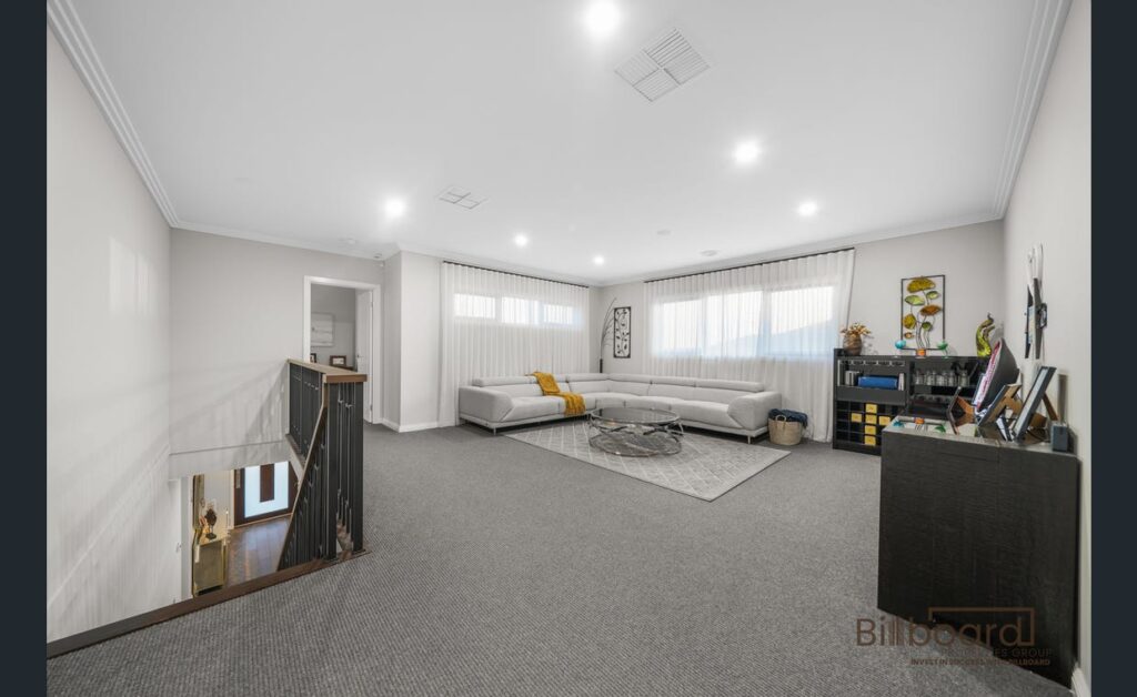 Spacious upstairs living/retreat area with grey carpet flooring, large corner sofa, coffee table on a rug, storage cabinet with decor pieces, wide windows with sheer curtains, and an open staircase leading down to the lower level.