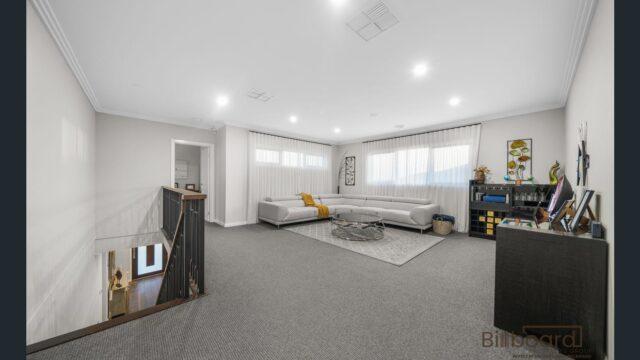 Spacious upstairs living/retreat area with grey carpet flooring, large corner sofa, coffee table on a rug, storage cabinet with decor pieces, wide windows with sheer curtains, and an open staircase leading down to the lower level.