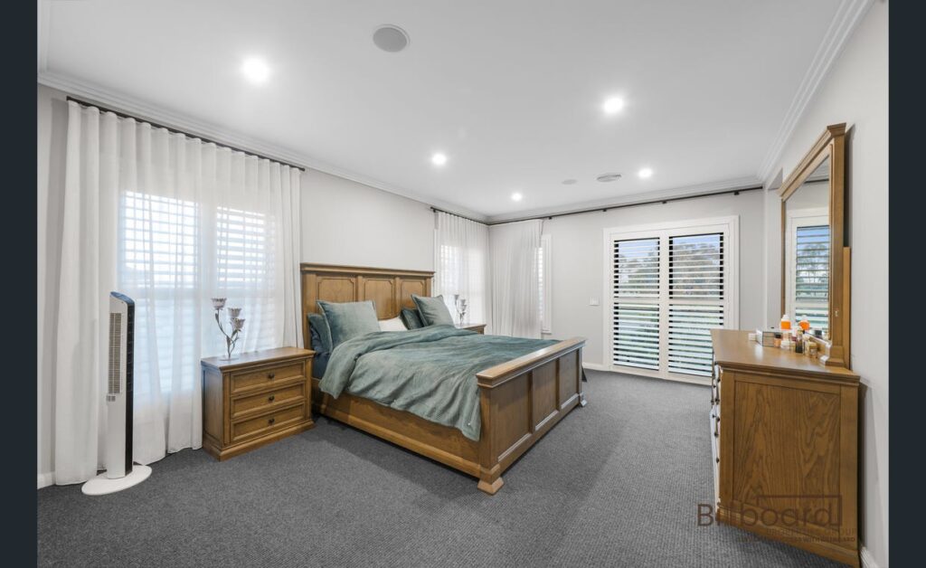 Large master bedroom featuring wooden bed frame and matching bedside tables, soft grey carpet flooring, sheer white curtains with plantation shutters, and bright recessed lighting creating a cozy and elegant interior setup.