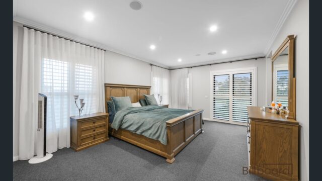Large master bedroom featuring wooden bed frame and matching bedside tables, soft grey carpet flooring, sheer white curtains with plantation shutters, and bright recessed lighting creating a cozy and elegant interior setup.