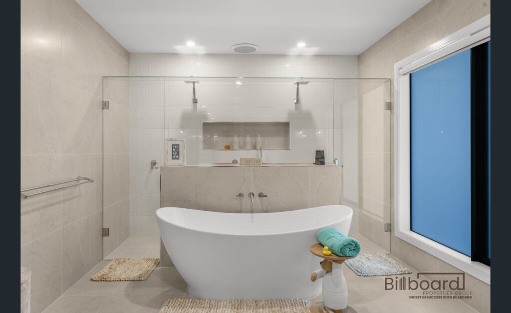 Luxurious modern bathroom with freestanding white bathtub placed centrally, large walk-in shower behind a glass screen, built-in wall niche with toiletries, soft neutral floor-to-ceiling tiles and a frosted window providing privacy and natural light.