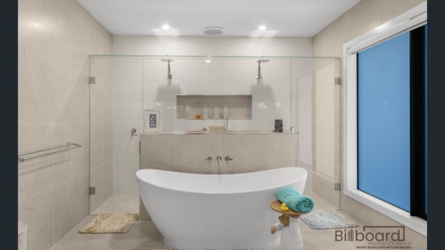 Luxurious modern bathroom with freestanding white bathtub placed centrally, large walk-in shower behind a glass screen, built-in wall niche with toiletries, soft neutral floor-to-ceiling tiles and a frosted window providing privacy and natural light.