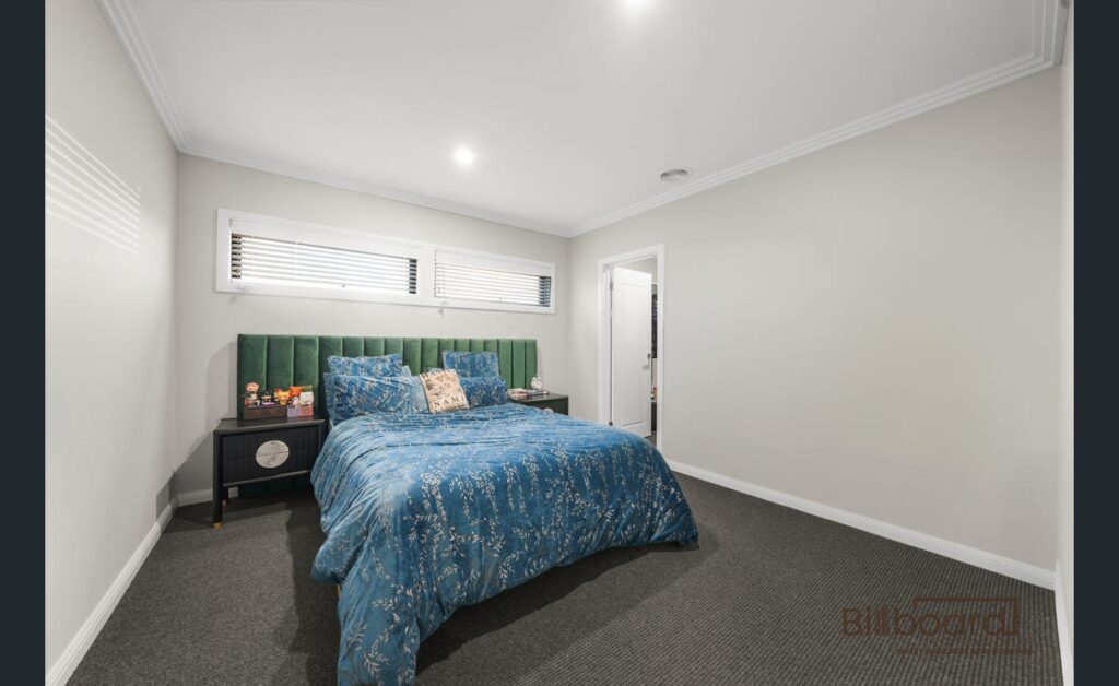 Bedroom with carpet flooring, green upholstered headboard, blue patterned bedding, bedside table with decor items, and high horizontal windows bringing in natural light, creating a simple and modern interior look.