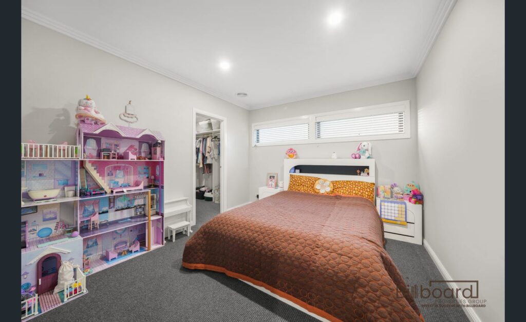 Kids bedroom with a neatly made single bed, brown quilt, large doll house and toys arranged along the wall, carpet flooring, and a walk-in robe visible through an open doorway. The room has a modern design with high windows letting in natural light.