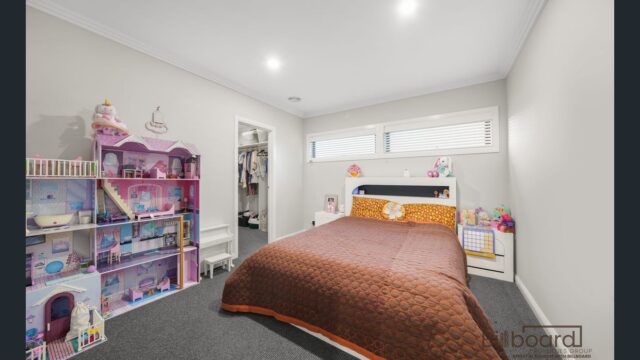 Kids bedroom with a neatly made single bed, brown quilt, large doll house and toys arranged along the wall, carpet flooring, and a walk-in robe visible through an open doorway. The room has a modern design with high windows letting in natural light.