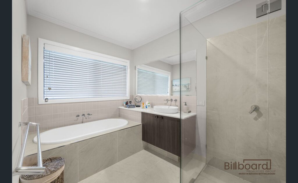 Bright bathroom featuring a built-in bathtub beneath a large window with blinds, modern vanity with mirror, neutral beige tiles, and a glass shower enclosure, presenting a clean and minimal contemporary design.