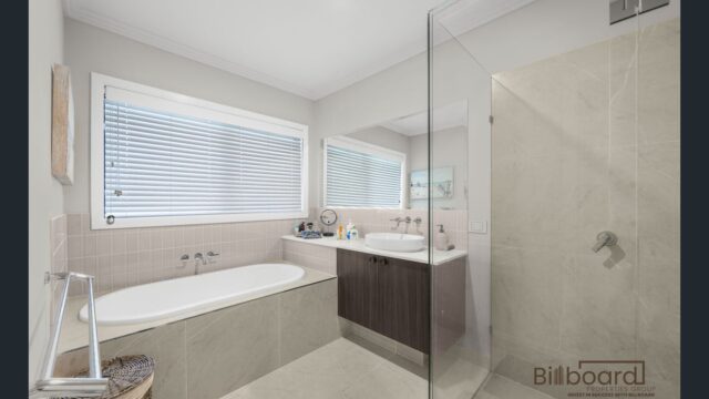 Bright bathroom featuring a built-in bathtub beneath a large window with blinds, modern vanity with mirror, neutral beige tiles, and a glass shower enclosure, presenting a clean and minimal contemporary design.