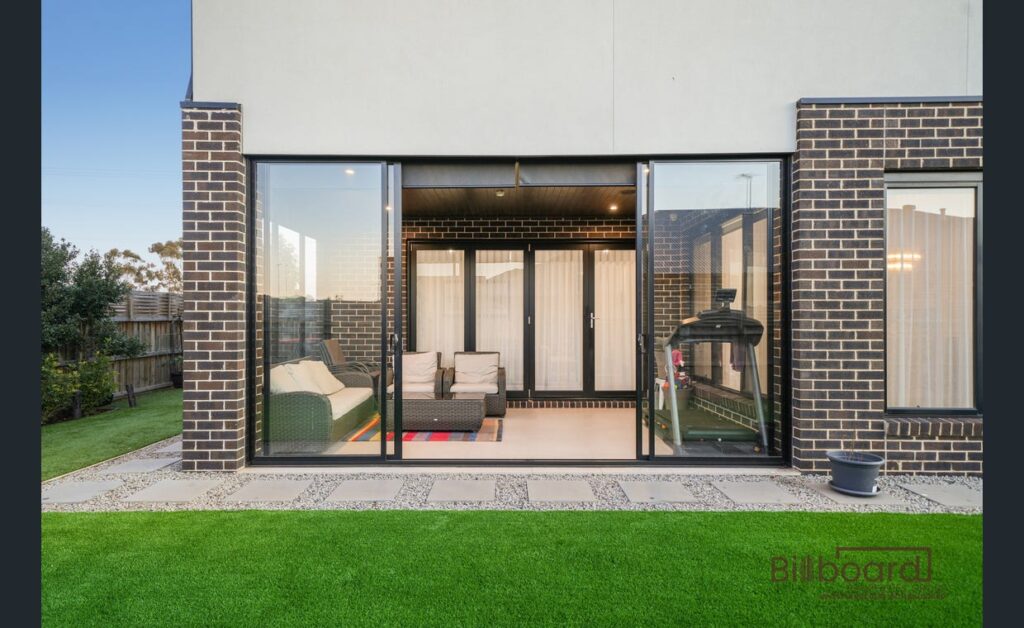 Outdoor view of a modern home with wide sliding glass doors opening into an indoor lounge seating area, surrounded by artificial lawn and a paved patio, with brick exterior walls framing the space.