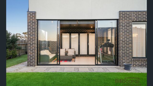 Outdoor view of a modern home with wide sliding glass doors opening into an indoor lounge seating area, surrounded by artificial lawn and a paved patio, with brick exterior walls framing the space.