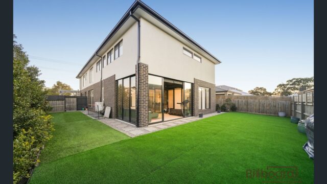 Backyard area with artificial lawn, paved walkway, potted plants along the fencing, BBQ placed near the timber fence, and a modern double-storey brick home on the right side with large sliding windows.