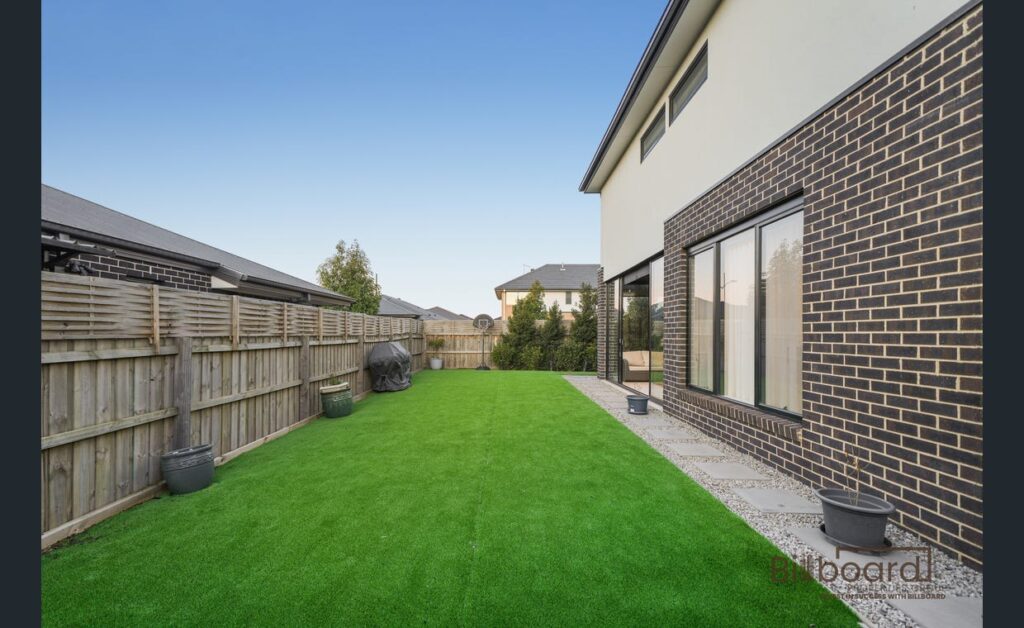 Backyard area with artificial lawn, paved walkway, potted plants along the fencing, BBQ placed near the timber fence, and a modern double-storey brick home on the right side with large sliding windows.