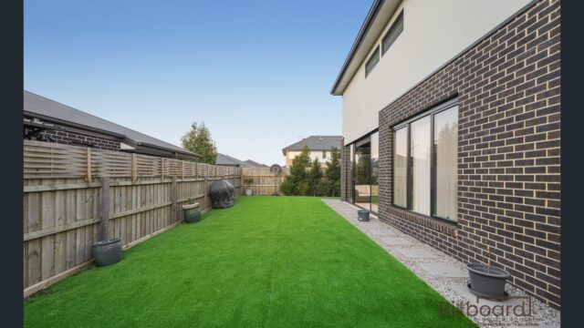Backyard area with artificial lawn, paved walkway, potted plants along the fencing, BBQ placed near the timber fence, and a modern double-storey brick home on the right side with large sliding windows.