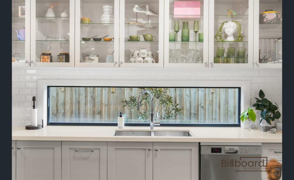 Close-up view of a modern kitchen sink area with white benchtop, stainless steel sink, cabinetry, dishwasher, glass display upper cabinets filled with neatly arranged dishes, and a long horizontal window looking out to the backyard fence.