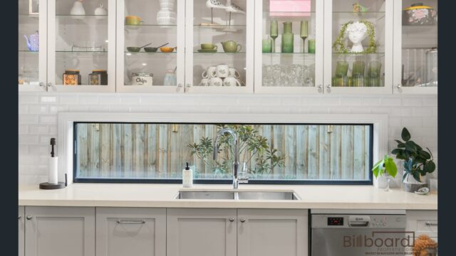 Close-up view of a modern kitchen sink area with white benchtop, stainless steel sink, cabinetry, dishwasher, glass display upper cabinets filled with neatly arranged dishes, and a long horizontal window looking out to the backyard fence.
