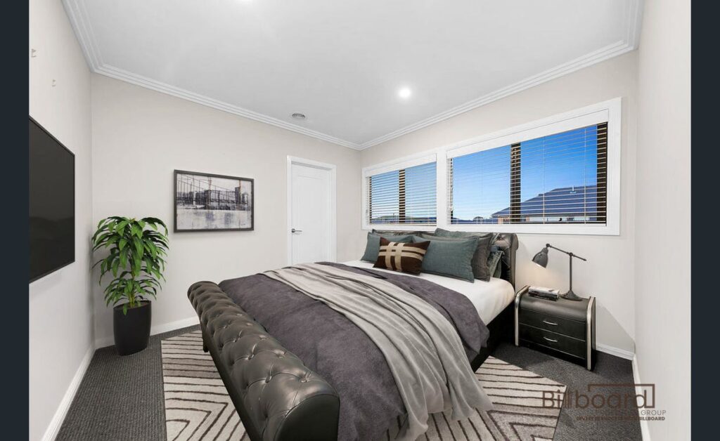 Modern bedroom with a neatly made bed styled in neutral grey bedding, patterned rug, black upholstered bench at the foot of the bed, bedside tables with lamps, wall art, indoor plant, and a wide window providing natural outdoor views.
