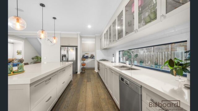 Modern kitchen with long white countertops, dishwasher, glass display upper cabinets, pendant lighting above the island, dark wooden flooring, and a long horizontal window splashback offering greenery views outside.