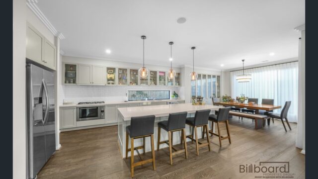 Open kitchen and dining area featuring a large island bench with bar seating, stainless steel appliances, white cabinetry, pendant lights, and a wooden dining table set near large windows with sheer curtains for natural light.