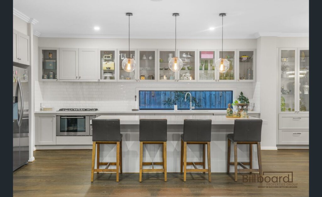 Bright modern kitchen with large island bench featuring four bar stools, white cabinetry, pendant lights hanging above the benchtop, built-in oven, gas cooktop, and long horizontal window splashback providing natural outdoor views.
