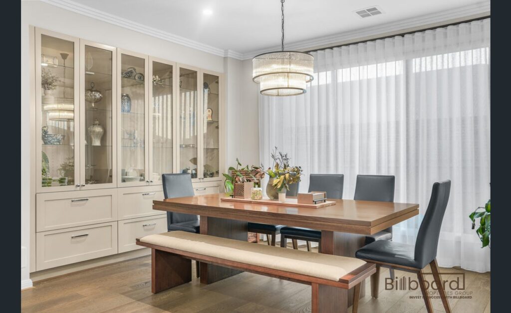 Elegant dining room with a wooden dining table, bench seating, cushioned dining chairs, ceiling chandelier light, large glass display cabinetry, and sheer curtains providing soft natural light into the space.
