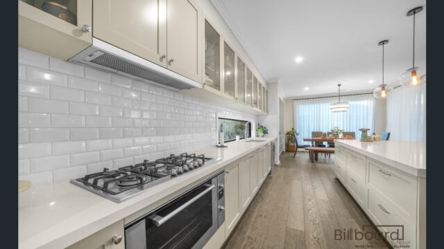 Sleek kitchen interior featuring a long benchtop with gas cooktop, subway tile backsplash, overhead display cabinets, built-in oven, and a large island bench with pendant lights, leading into an open dining and living space.