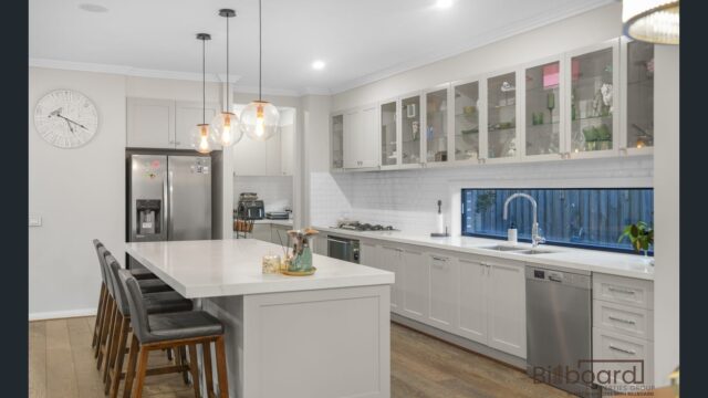 Modern kitchen with white cabinetry, large island bench with bar stools, pendant lighting, stainless steel appliances, gas cooktop, glass display upper cabinets and a long window splashback providing natural light.
