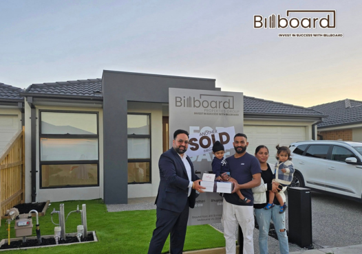 A family receiving house keys from a representative in front of a new home with a 'Sold' sign.