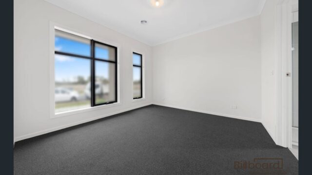 Spacious empty bedroom with grey carpet flooring, white walls, large windows with black frames allowing natural light, and a ceiling light fixture. The room has a clean, modern interior design ready for furnishing.