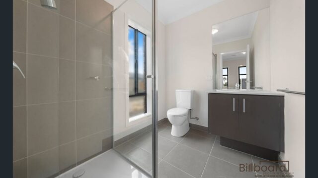 Modern bathroom interior featuring a glass shower enclosure, toilet, grey floor tiles, a wall-mounted dark vanity cabinet with a large mirror, and a tall window allowing natural daylight into the space. The room has a clean and minimal contemporary design.