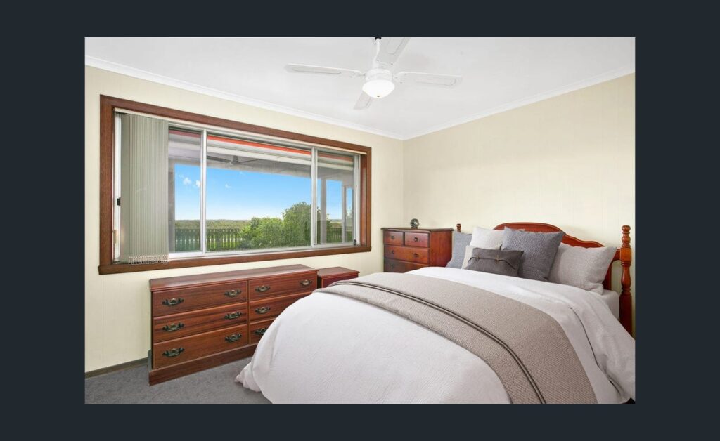 Cozy farmhouse style bedroom with wooden furniture, ceiling fan and large window overlooking green countryside views.