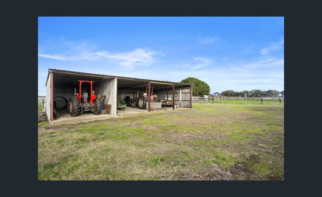 Open farm machinery shed housing tractors on a large rural property with green open land and clear blue sky.