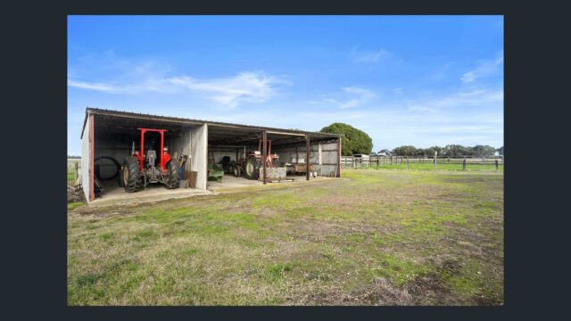 Open farm machinery shed housing tractors on a large rural property with green open land and clear blue sky.