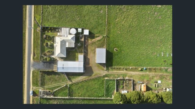Aerial top view of rural farm property showing house, sheds, fenced paddocks and surrounding green farmland.