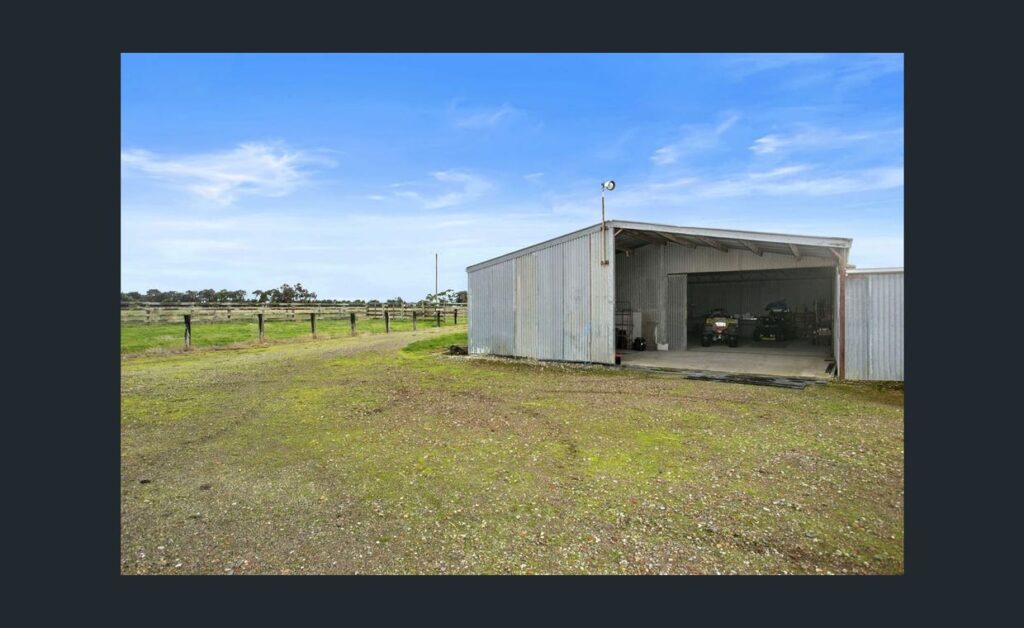 Open farm shed on rural property with machinery inside and large cleared land area under a blue sky.
