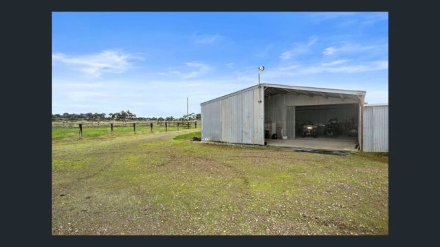 Open farm shed on rural property with machinery inside and large cleared land area under a blue sky.
