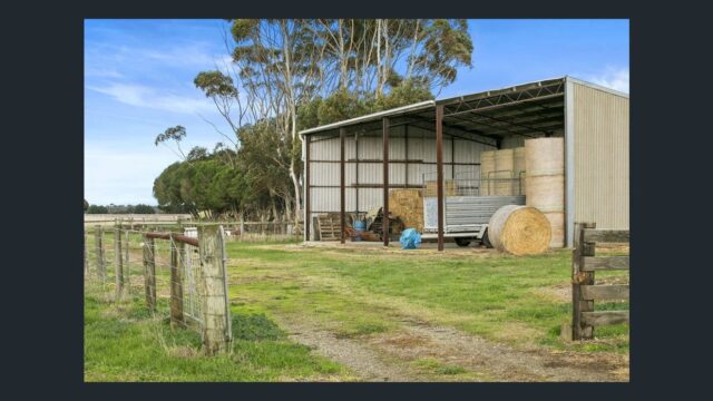 Farm storage shed with hay bales, machinery and fencing on a rural agricultural property surrounded by open land and trees.
